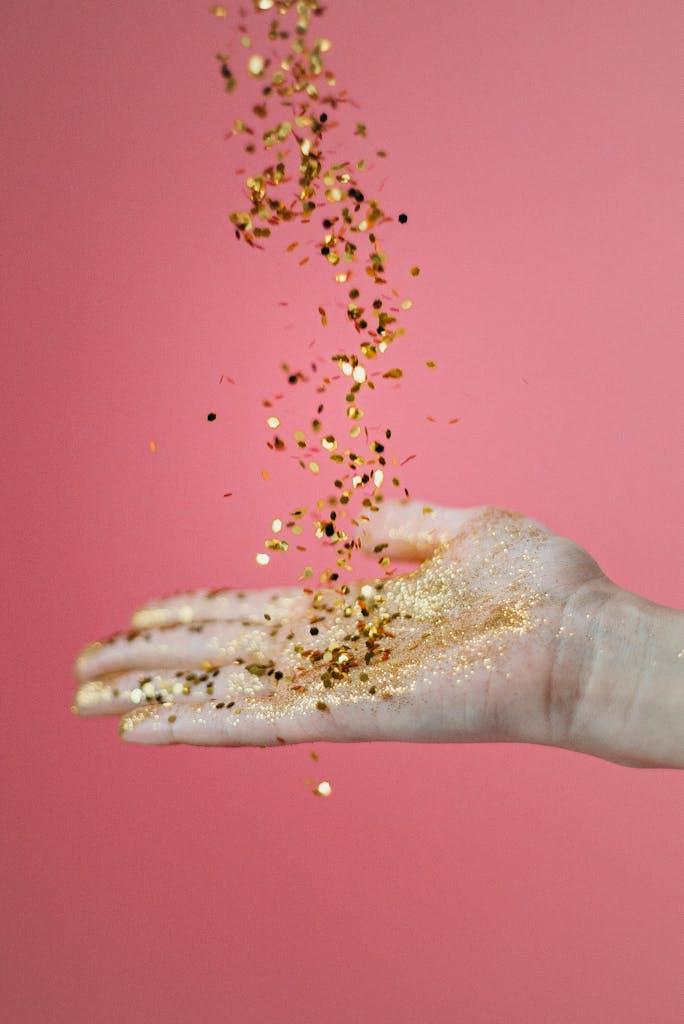 A cascade of gold confetti pours onto an open hand against a pink background.