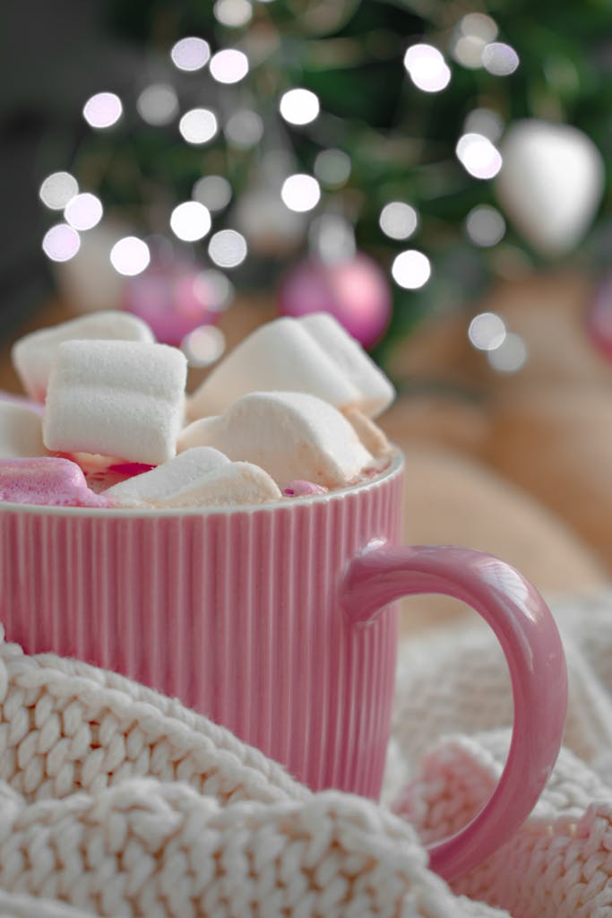 A cozy pink mug of hot cocoa with marshmallows, set against a festive bokeh background.